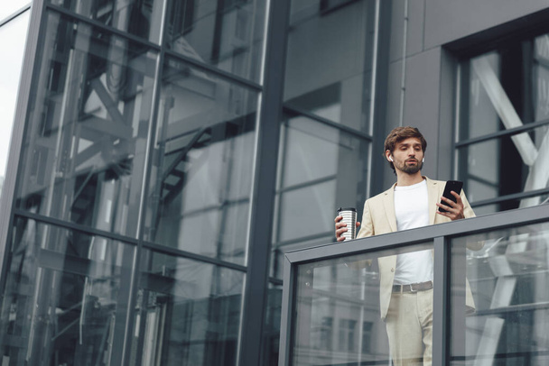 Retrato de un hombre serio vestido con traje de negocios de moda que tiene videollamada en el teléfono inteligente mientras está parado al aire libre con una taza de café. Empresario teniendo conversación de trabajo cerca de edificio de oficinas. - Foto, Imagen