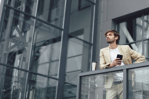 Молодий бородатий бізнесмен в модному костюмі, стоїть на балконі власного офісу і використовує смартфон. Красивий в навушниках чоловік працює на свіжому повітрі з чашкою кави
. - Фото, зображення