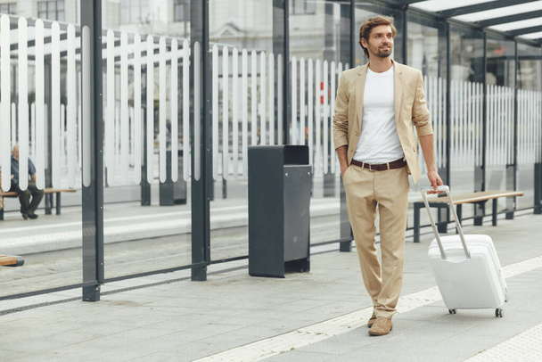 Stylový mladý muž s plnovousem drží bílý kufr, zatímco stojí na autobusové zastávce. Hezký podnikatel čeká na vozidlo venku. - Fotografie, Obrázek