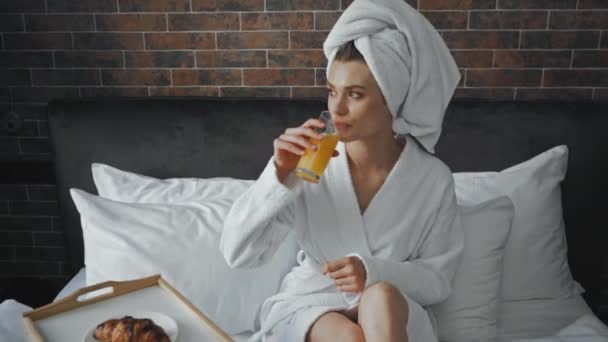 giovane donna in asciugamano bere succo d'arancia vicino vassoio con croissant  - Filmati, video