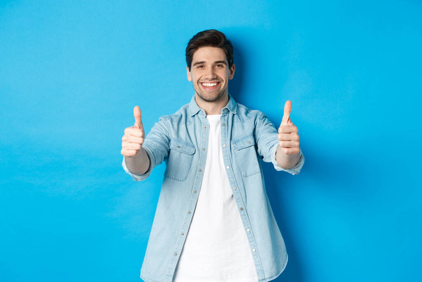 Glimlachende zelfverzekerde man die duimen laat zien, kwaliteit garandeert, iets goeds goedkeurt, tegen een blauwe achtergrond staat - Foto, afbeelding