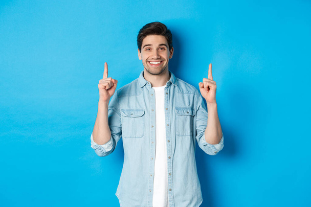 Porträt eines gutaussehenden bärtigen Mannes in lässigem Outfit, glücklich lächelnd und mit erhobenem Finger auf den Kopierraum zeigend, vor blauem Hintergrund stehend - Foto, Bild