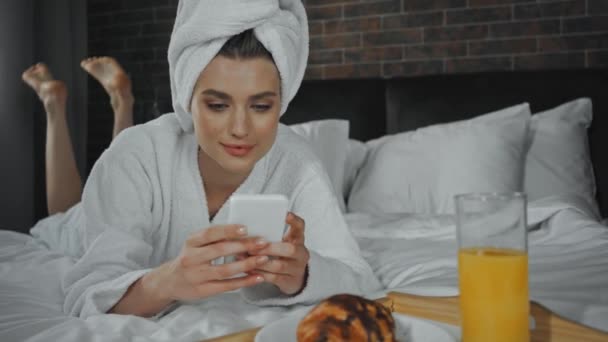 mulher alegre em toalha deitada na cama e usando smartphone perto do café da manhã - Filmagem, Vídeo