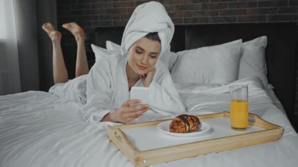 młoda kobieta w ręczniku leżąca na łóżku i korzystająca ze smartfona przy śniadaniu - Materiał filmowy, wideo