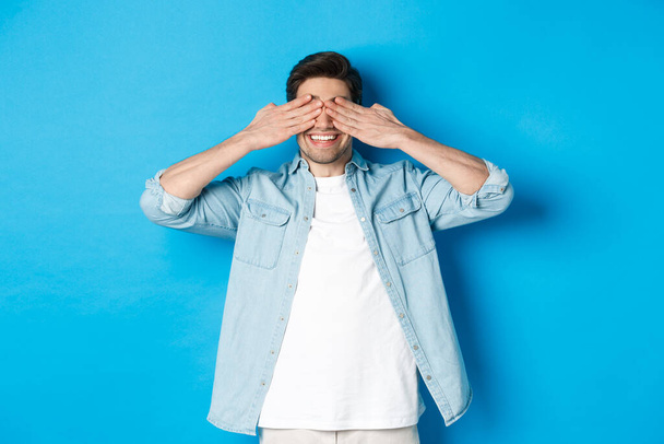 Smiling adult man waiting for surprise, covering eyes with hands and anticipating, standing against blue background in casual clothes - Photo, Image