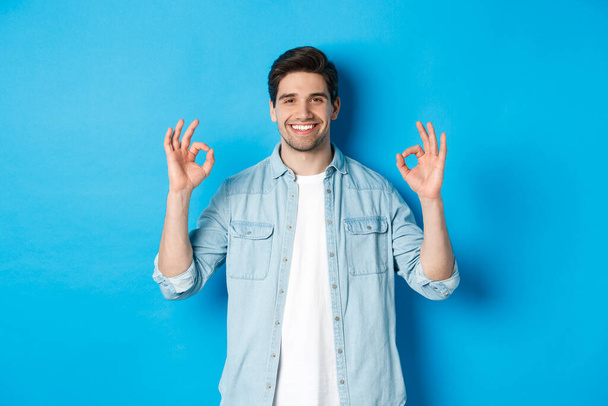 Уверенный в себе улыбающийся взрослый мужчина, показывающий в порядке признаки и выглядящий довольным, как что-то, стоя на синем фоне - Фото, изображение