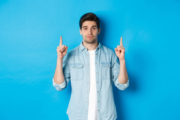 Kép nyugodt jóképű férfi mutatja promo ajánlat, ujjal felfelé a másolás tér, álló ellen kék háttér - Fotó, kép