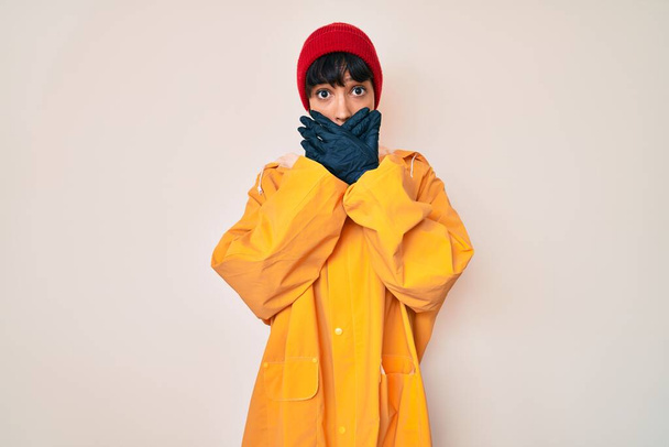 Beautiful brunettte woman wearing yellow raincoat shocked covering mouth with hands for mistake. secret concept.  - Foto, Bild