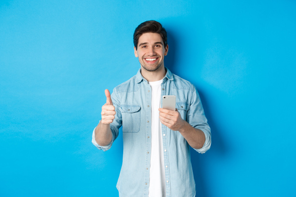 Fogalom az online vásárlás, alkalmazások és technológia. Elégedett férfi alkalmi ruhák mosolygós, mutatja hüvelykujját fel használata után smartphone app, álló felett kék háttér - Fotó, kép