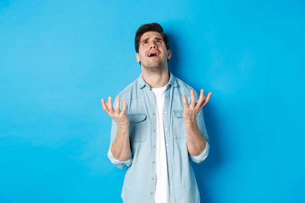 Frustrated guy complaining to god, looking up and pleading, standing against blue background upset - Photo, Image
