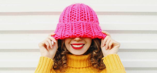 Winter portrait close up of cheerful woman pulling pink hat over her eyes wearing a yellow knitted sweater on white background - Φωτογραφία, εικόνα