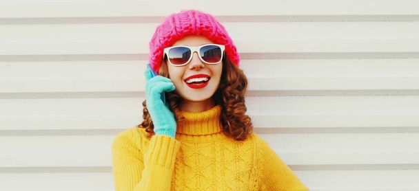 Portrait of happy smiling young woman calling on smartphone wearing a colorful pink knitted hat, yellow sweater over white background - 写真・画像