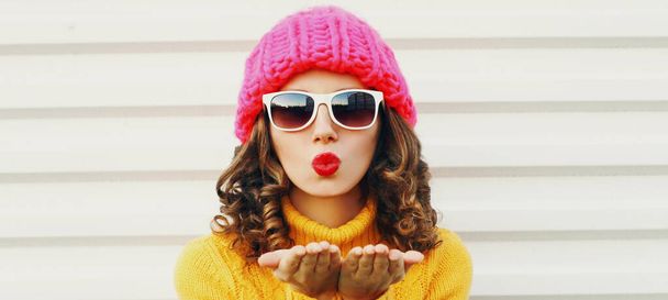 Retrato de invierno primer plano de la joven mujer soplando labios rojos enviando beso de aire dulce con un sombrero rosa, suéter amarillo sobre fondo blanco - Foto, Imagen