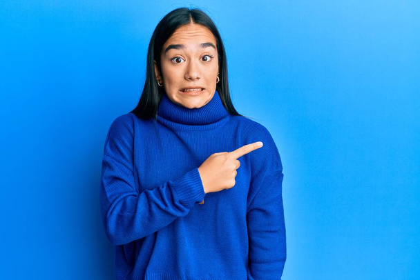 Молодая азиатская женщина в обычном зимнем свитере, указывая в сторону взволнованный и нервный указательным пальцем, обеспокоенный и удивленный выражение  - Фото, изображение