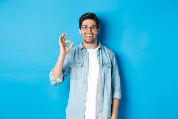 Jeune homme attrayant portant des lunettes et des vêtements décontractés, montrant ok bon signe dans l'approbation, comme quelque chose, debout sur fond bleu - Photo, image