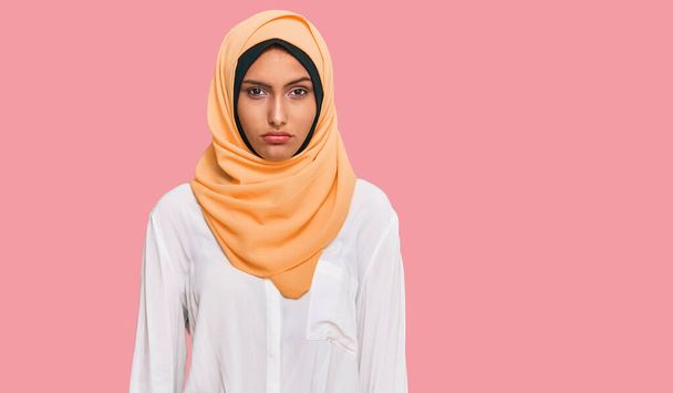 Genç esmer kadın geleneksel İslami başörtüsü takıyor ve yüzünde ciddi bir ifade var. Basit ve doğal bir şekilde kameraya bakıyorsun..  - Fotoğraf, Görsel