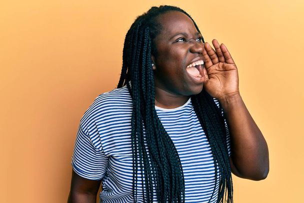 Mujer negra joven con trenzas que usan ropa casual gritando y gritando fuerte a un lado con la mano en la boca. concepto de comunicación.  - Foto, imagen