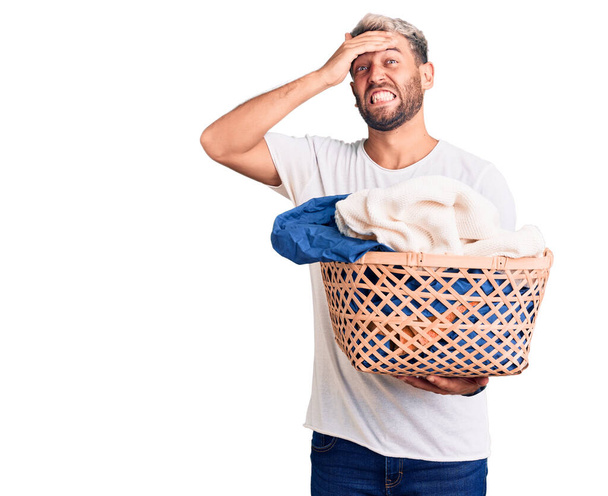Joven hombre rubio guapo sosteniendo cesta de la ropa con ropa estresada y frustrada con la mano en la cabeza, cara sorprendida y enojada  - Foto, imagen