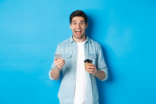Ευτυχισμένος νεαρός άνδρας πίνοντας καφέ και χρησιμοποιώντας το κινητό τηλέφωνο, αναζητούν ενθουσιασμένος, στέκεται πάνω σε μπλε φόντο - Φωτογραφία, εικόνα