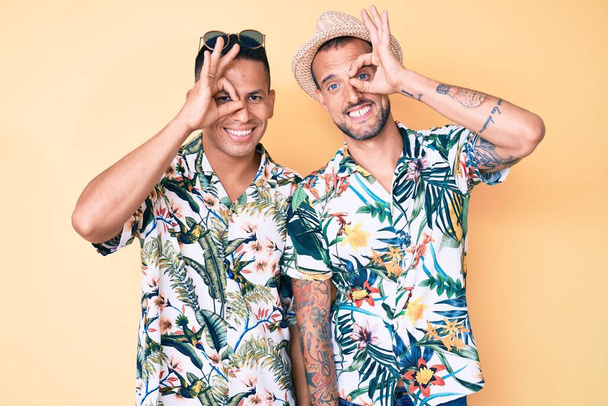 Young gay couple of two men wearing summer hat and hawaiian shirt doing ok gesture with hand smiling, eye looking through fingers with happy face.  - Foto, immagini