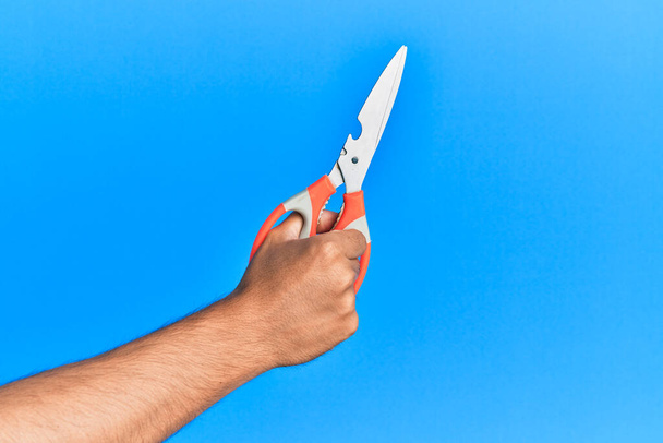 Рука молодого іспаномовного чоловіка, використовуючи ножиці над ізольованим синім тлом
. - Фото, зображення