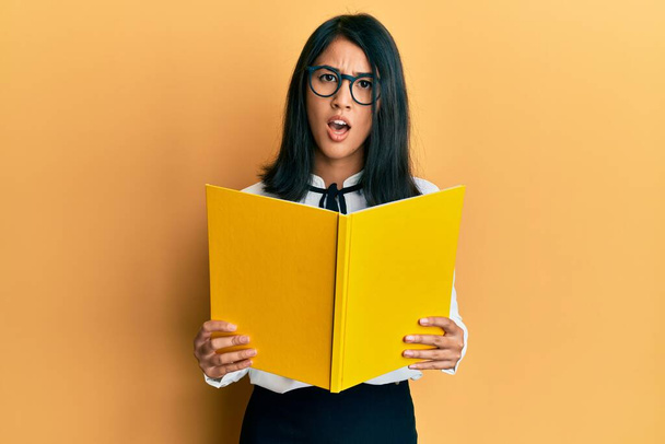 Piękna azjatycka młoda kobieta czytająca książkę noszącą okulary w szoku na twarzy, wyglądająca sceptycznie i sarkastycznie, zaskoczona otwartymi ustami  - Zdjęcie, obraz