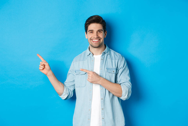 Bello uomo caucasico in abito casual, puntando il dito verso sinistra e sorridente, mostrando offerta promozionale, in piedi su sfondo blu - Foto, immagini
