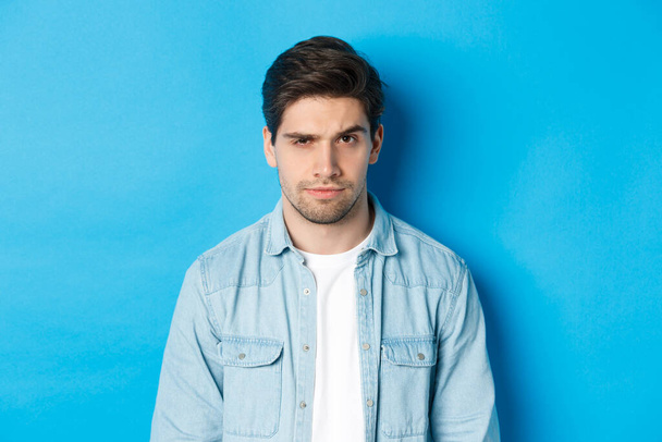 Close-up of suspicious and serious guy raising one eyebrow, looking at camera, standing against blue background - Fotoğraf, Görsel