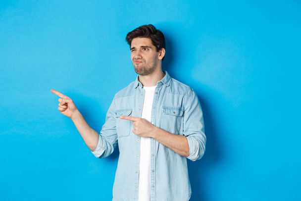 Doubtful adult man pointing fingers left at promotion and looking unsure, grimacing disappointed, standing against blue background - Photo, image