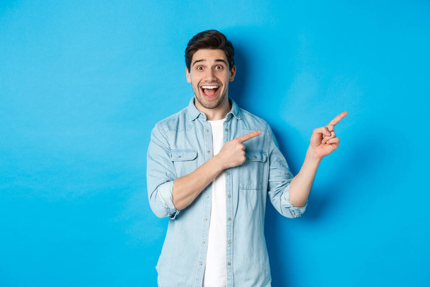 Image of excited handsome man in casual outfit, showing advertisement, pointing fingers right at copy space and smiling, standing against blue background - 写真・画像