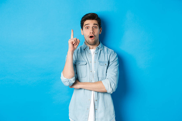 Image of handsome adult man having an idea, raising finger and smiling excited, found solution, standing against blue background - Fotoğraf, Görsel
