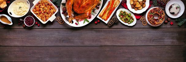 Classic Christmas turkey dinner. Overhead view top border on a dark wood banner background with copy space. Turkey, potatoes and sides, dressing, fruit cake and plum pudding. - Photo, Image