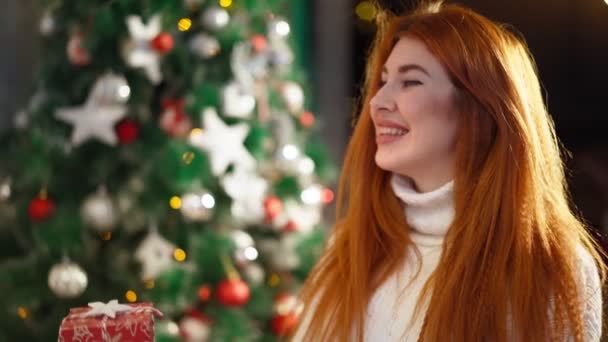 Όμορφη ευτυχισμένη κοκκινομάλλα γυναίκα κάθεται φόντο Χριστουγεννιάτικο δέντρο και χαμογελώντας - Πλάνα, βίντεο