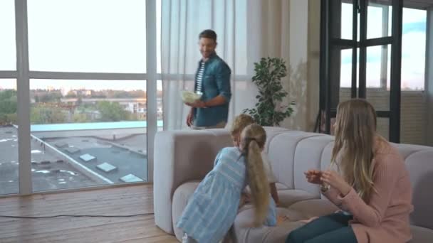 Cheerful father giving bowl with popcorn for family on couch in living room - Imágenes, Vídeo