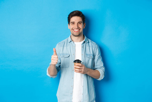 Komea nuori mies näyttää peukaloa ja juo kahvia, suosittelee kahvilan noutoa, seisoo sinisen taustan yläpuolella - Valokuva, kuva