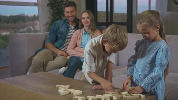 Αδέρφια παίζουν μπλοκ παιχνίδι ξύλο με ευτυχείς γονείς στο παρασκήνιο στο σπίτι - Πλάνα, βίντεο