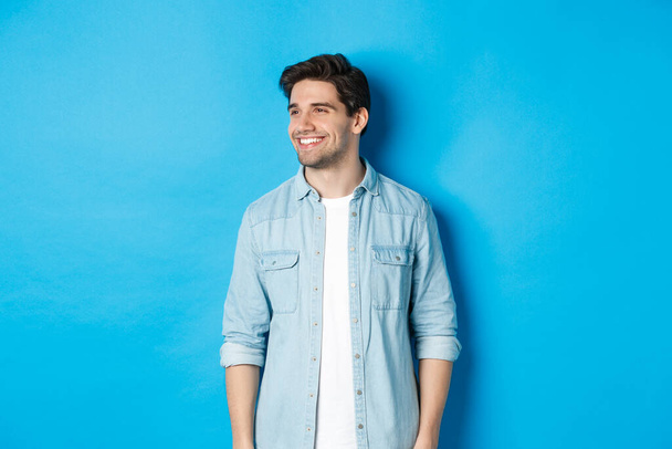 Beau souriant homme adulte en tenue décontractée, souriant et regardant à gauche à l'offre promo, debout sur fond bleu - Photo, image