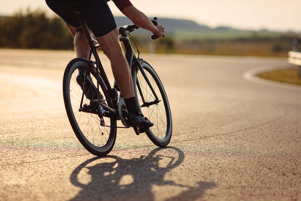 Primer plano del ciclista profesional de carretera con piernas fuertes en bicicleta por carretera pavimentada durante el tiempo de la mañana. Concepto de estilo de vida activo y saludable. - Foto, Imagen