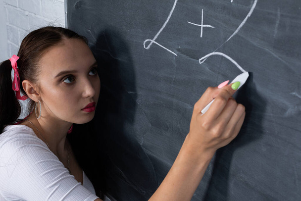 Teenager chalks a math equation on a dark gray school blackboard. Her hair is pinned with a pink ribbon in two ponytails. - Foto, Bild