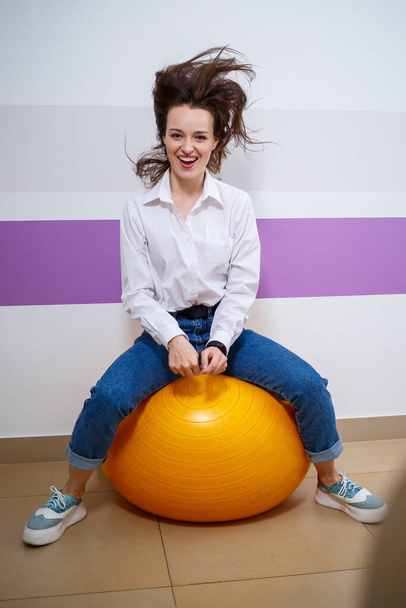 Una chica de apariencia europea con diferentes emociones en su cara se sienta en una pelota para una gimnasta. Atractiva joven morena sonriendo y jugando - Foto, imagen