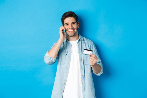 Handsome man calling bank and holding credit card, having mobile conversation, standing over blue background - Photo, Image