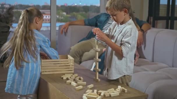 Hermanos jugando bloques juego de madera con los padres sentados en el sofá en el fondo - Imágenes, Vídeo