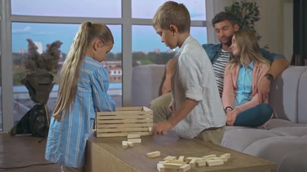 Hermanos embalaje bloques juego de madera en caja con los padres en el fondo en casa - Metraje, vídeo