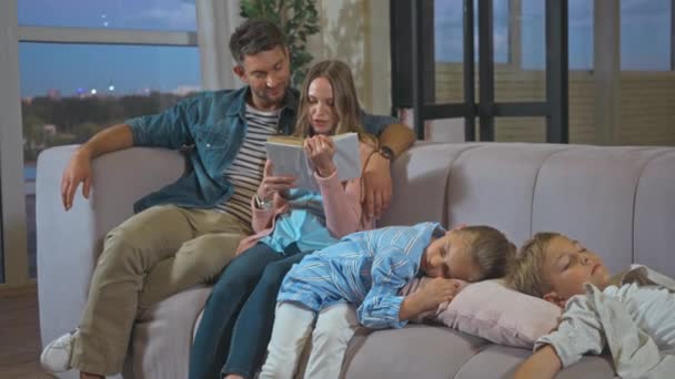 Mother reading book near children sleeping on couch in living room - Footage, Video