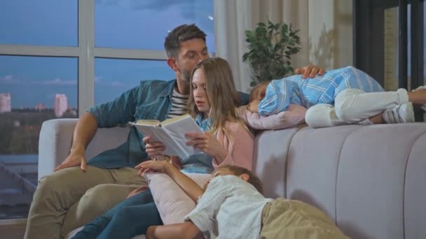 Padre y madre con el libro sentado cerca de los niños durmiendo en el sofá en casa - Metraje, vídeo