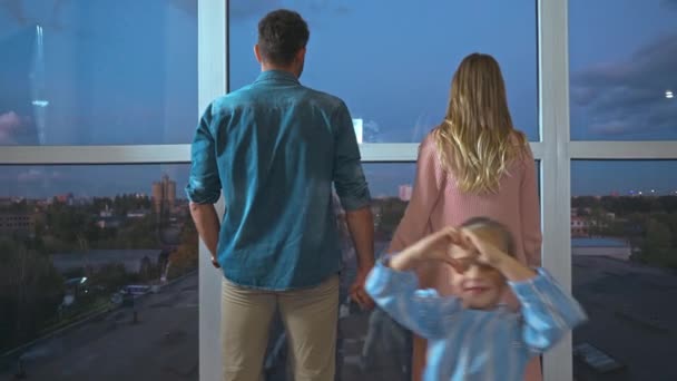 Szczęśliwa córka pokazuje gest miłości w pobliżu rodziców trzymających się za ręce przy oknach - Materiał filmowy, wideo