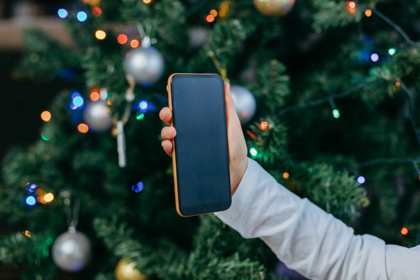 Мобільний телефон в руках дівчини на тлі новорічної ялинки. Онлайн привітання на Новий рік. Інтернет покупки
. - Фото, зображення