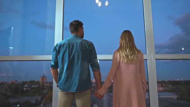 Vista trasera de pareja romántica cogida de la mano cerca de ventanas panorámicas en casa - Metraje, vídeo