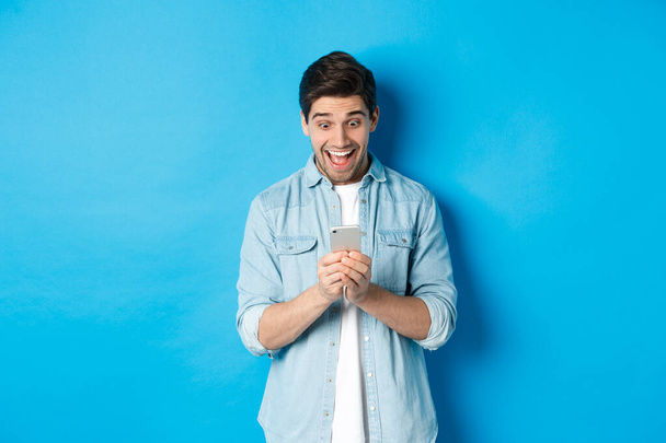 Bild eines aufgeregten Mannes, der auf sein Handy lächelt, auf dem Smartphone online einkauft und vor blauem Hintergrund steht - Foto, Bild