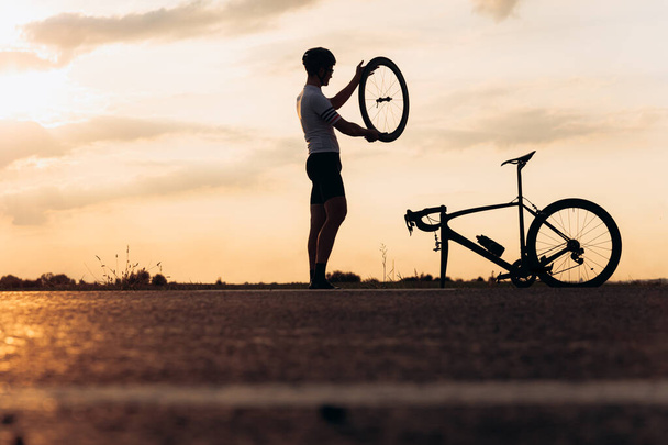 Silhouette di uomo attivo in silhouette riparazione bici su strada asfaltata. YOung ragazzo in abbigliamento sportivo e casco ruota di fissaggio dopo la sera in bicicletta. - Foto, immagini